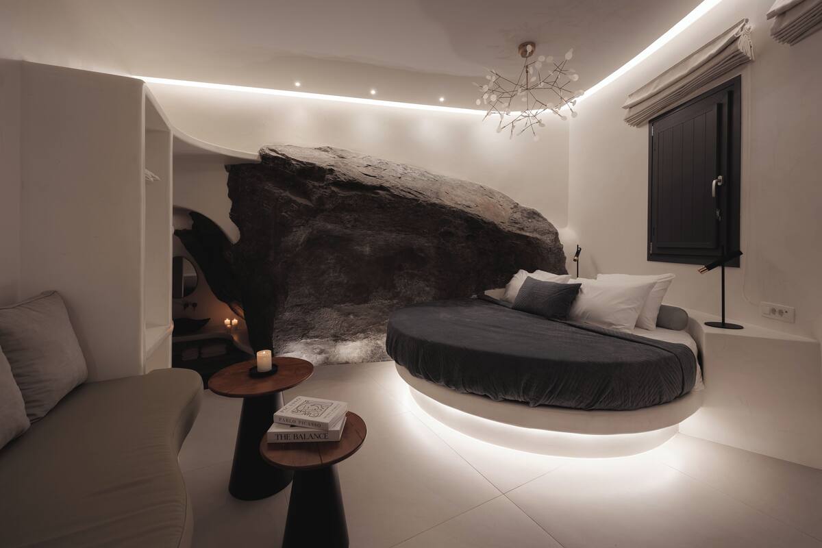 Un dormitorio moderno con una gran formación rocosa, una cama curvada y una mesita con un libro.