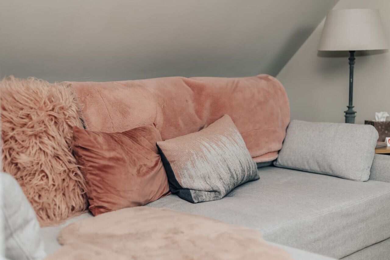 A grey sofa with pink and grey cushions, a fluffy pink throw, and a lamp on a bedside table.