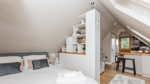 Une chambre moderne avec un grand lit, une armoire blanche et un coin cuisine visible à travers une porte ouverte.