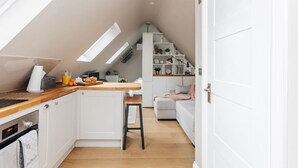 Une cuisine moderne avec un plan de travail en bois, des armoires blanches et un four encastré. On y trouve un coin repas avec une table et des chaises blanches, ainsi qu’un escalier menant à une mezzanine aménagée en espace nuit.