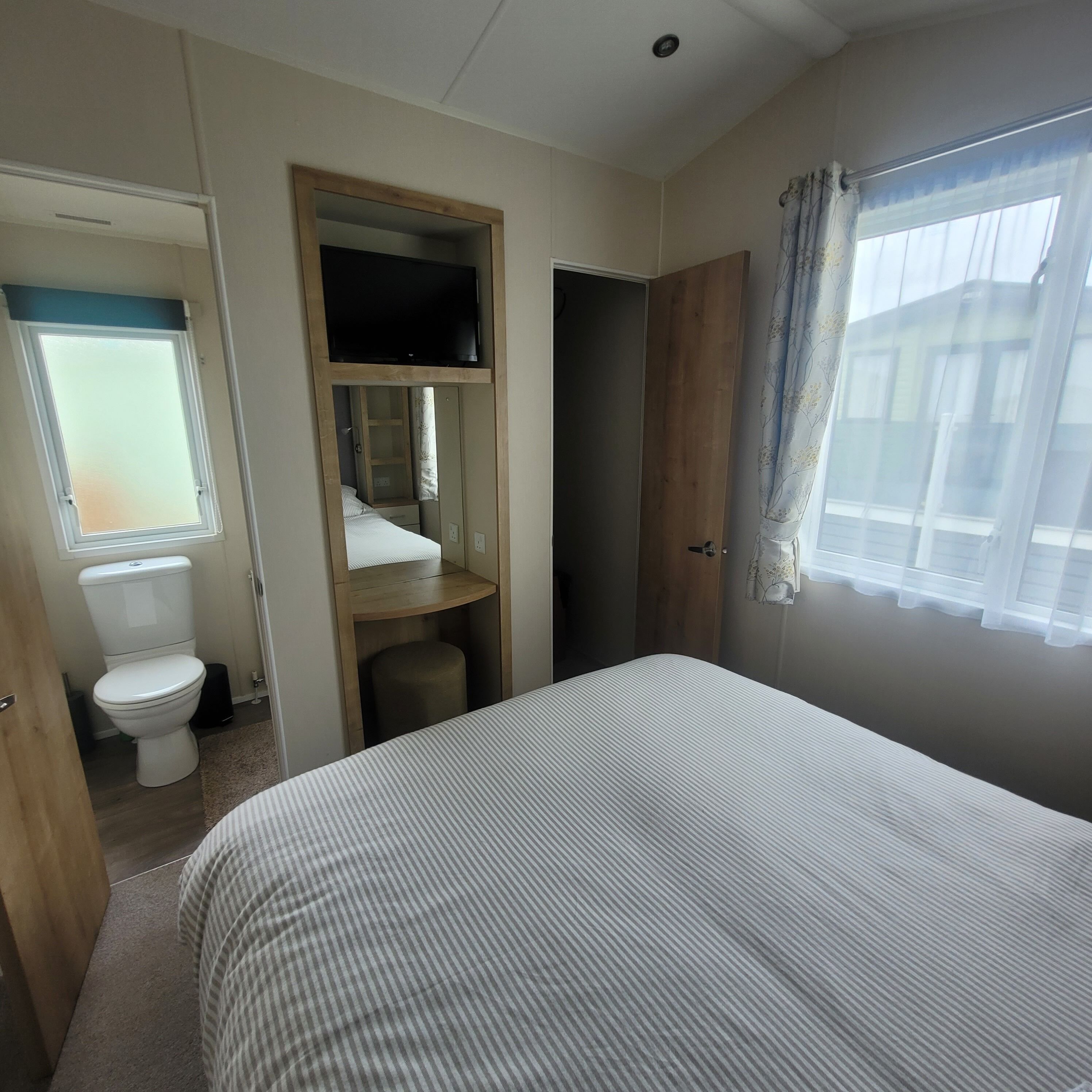 A bedroom with a bed, a wooden wardrobe, a television, a toilet, and a window with curtains.