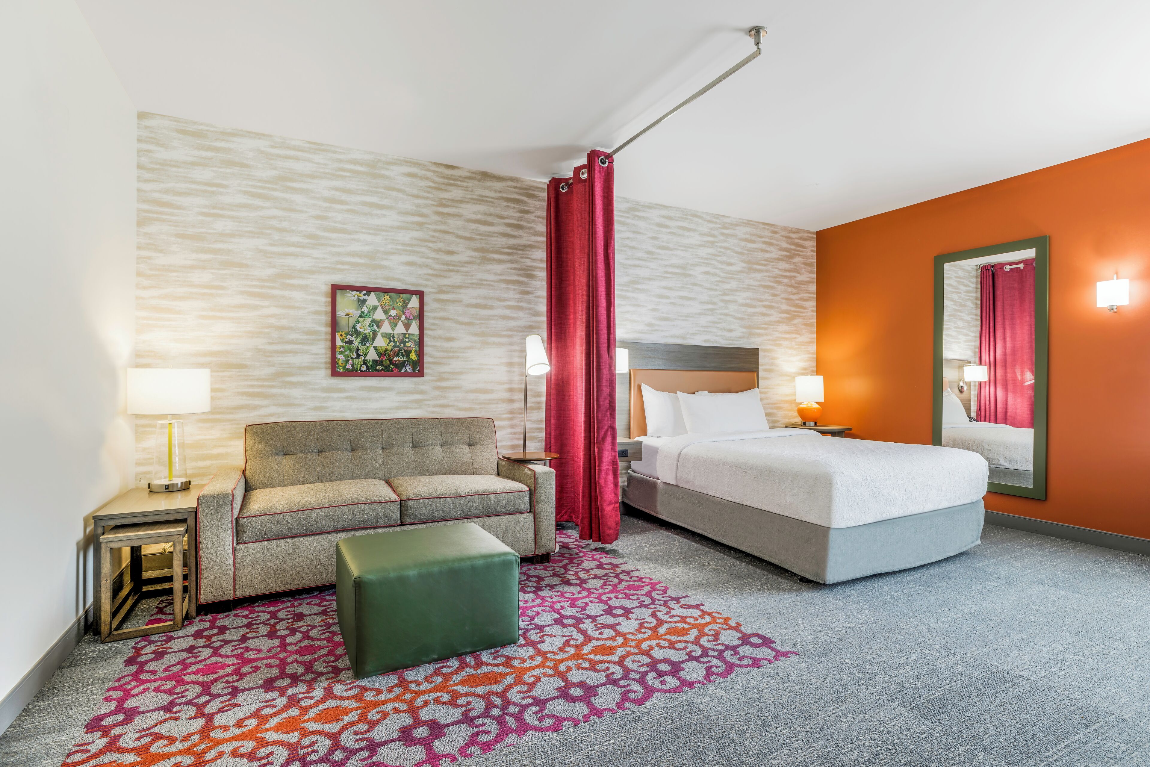 A hotel room with a bed, a sofa, a small table, a lamp, a decorative rug, and a mirror.