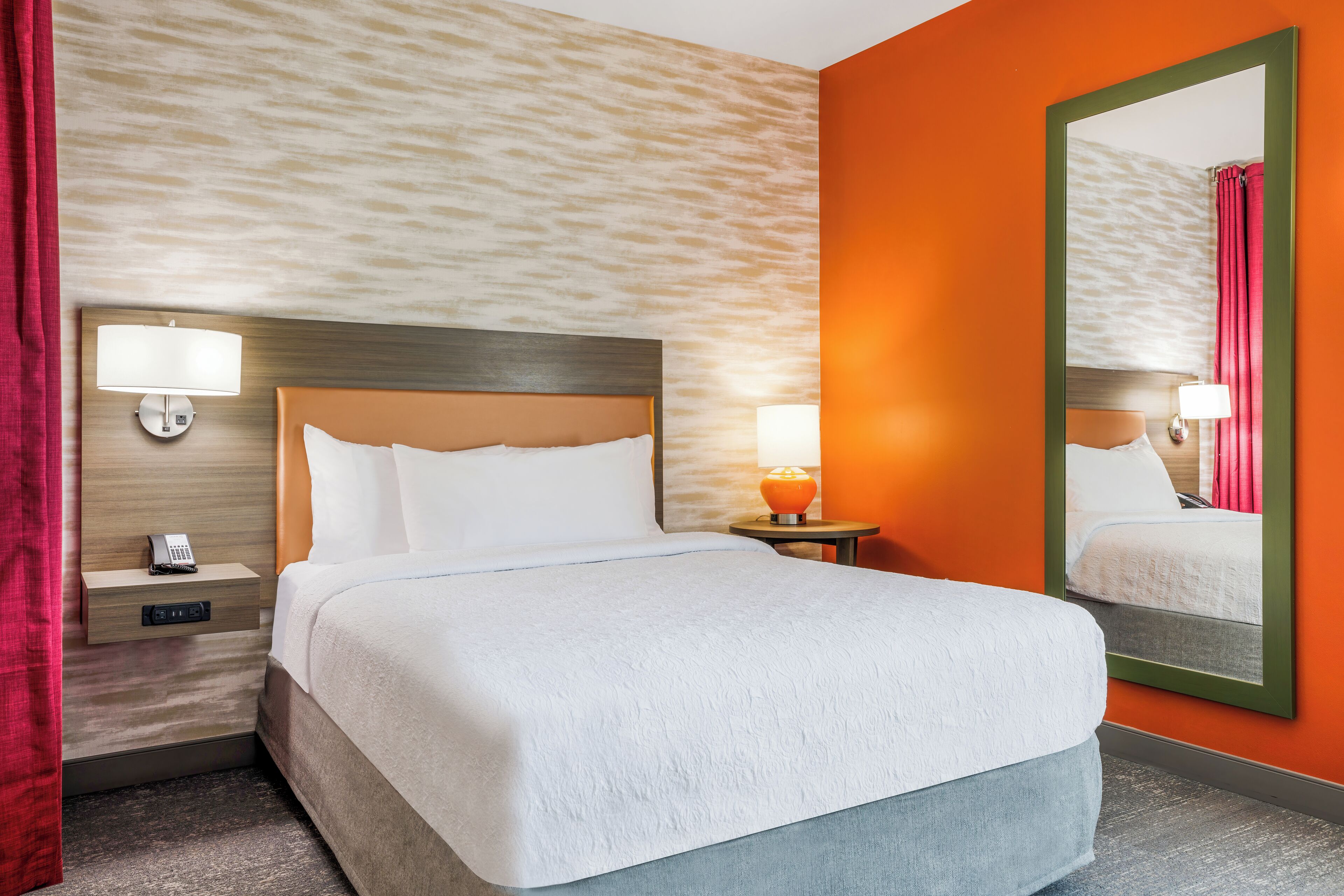A hotel room with a bed, nightstand, lamp, and mirror.