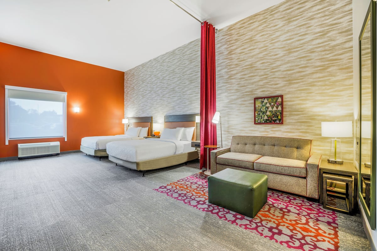 A hotel room with two beds, a sofa, a green ottoman, and a red curtain.