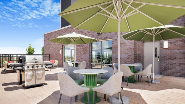 An outdoor patio with round tables and chairs, green umbrellas, and a barbecue grill. - Home2 Suites Hilton Aurora Medical Center (Aurora)