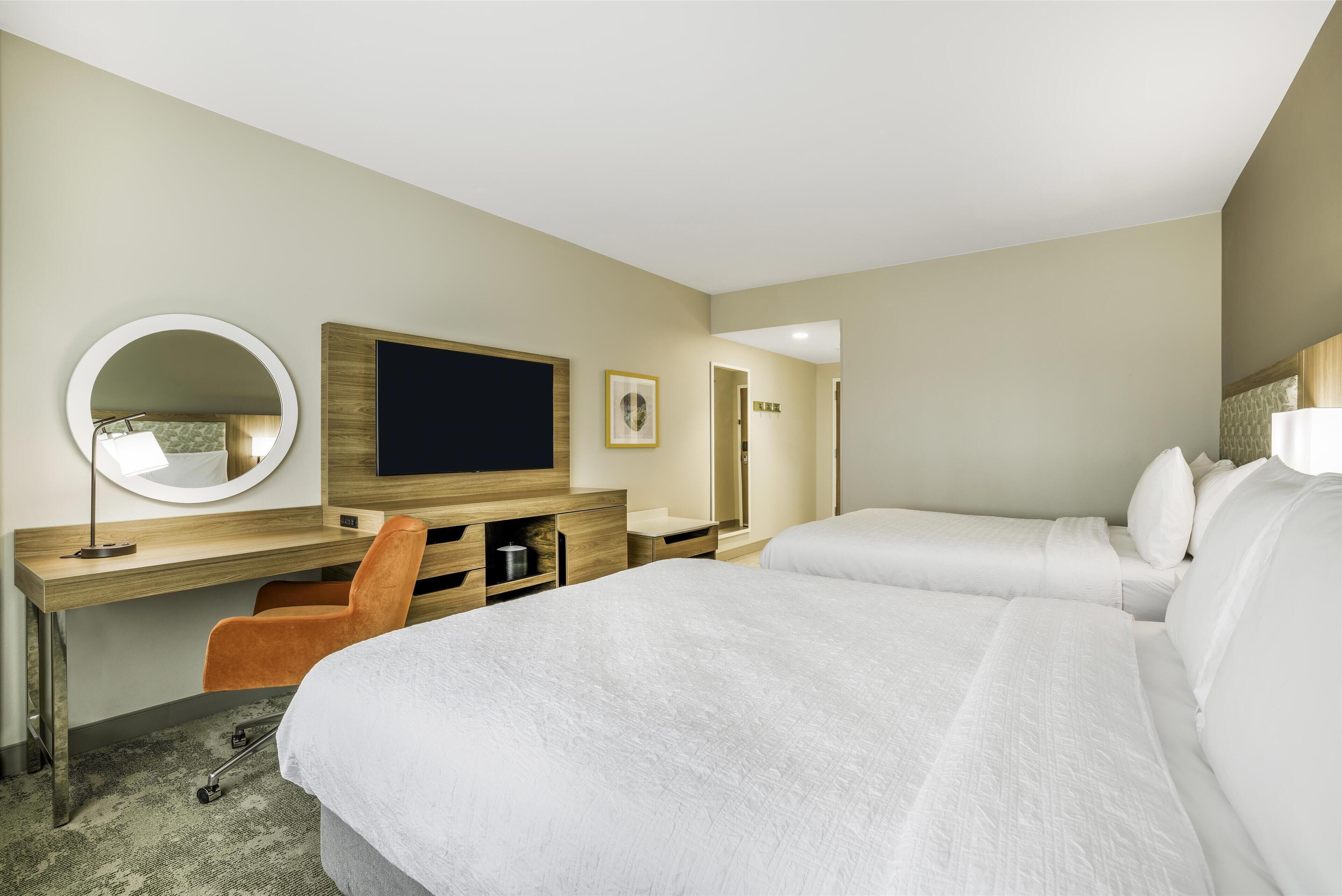 Quarto de hotel com uma cama, uma escrivaninha com cadeira, televisão, espelho e guarda-roupa.