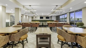 A modern dining area with multiple tables and chairs, large windows offering a view, and a central seating area with a green couch. - Home2 Suites Hilton Aurora Medical Center (Aurora)