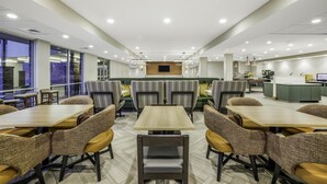 A spacious dining area with multiple tables and chairs, a central seating area with cushioned benches, and a kitchen in the background. - Home2 Suites Hilton Aurora Medical Center (Aurora)