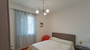 Un dormitorio con cama, mesita de noche, una silla roja, ventana con cortinas e iluminación en el techo.