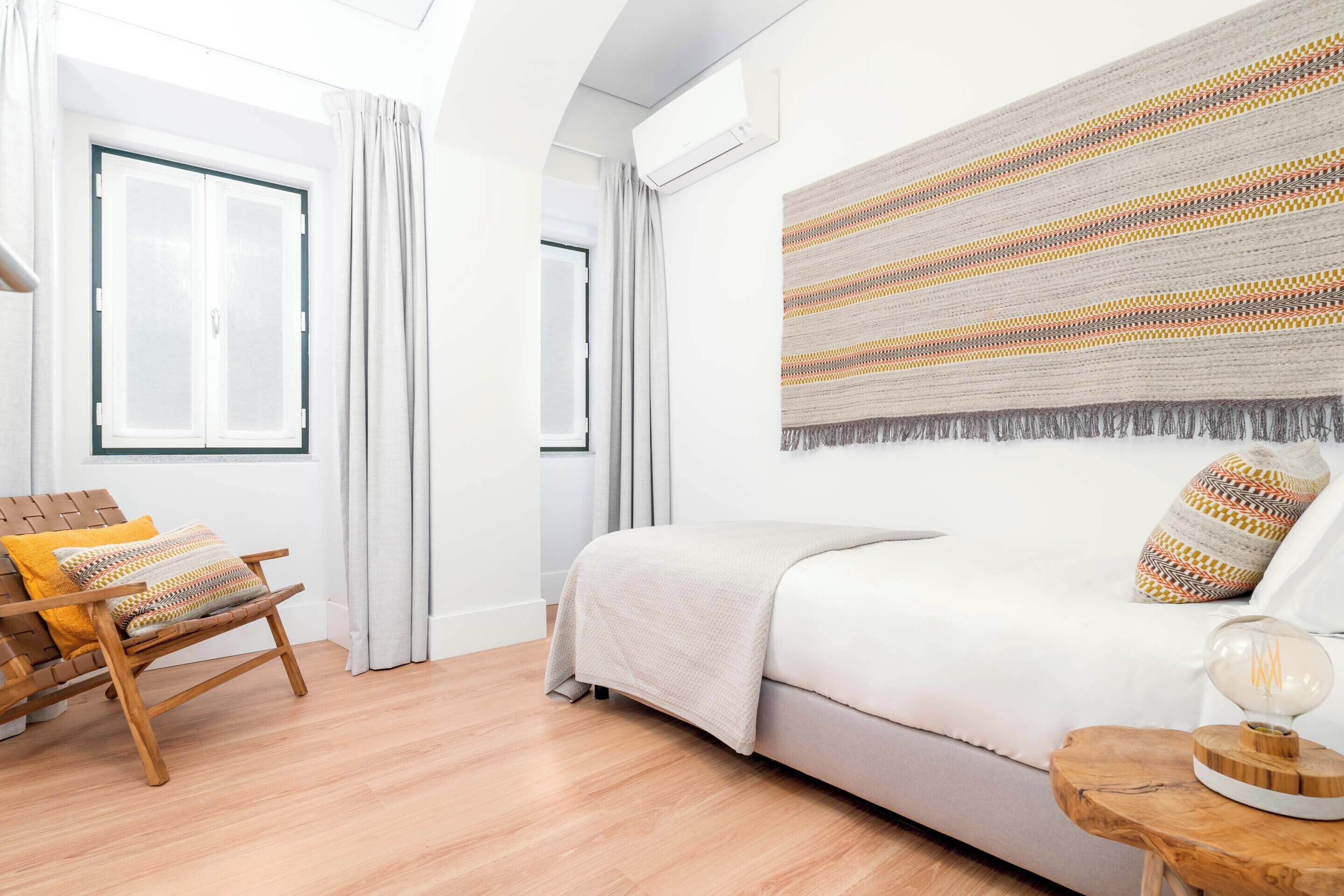 Une chambre moderne avec un lit, une chaise en bois, un tapis à motifs et un luminaire fixé au mur.