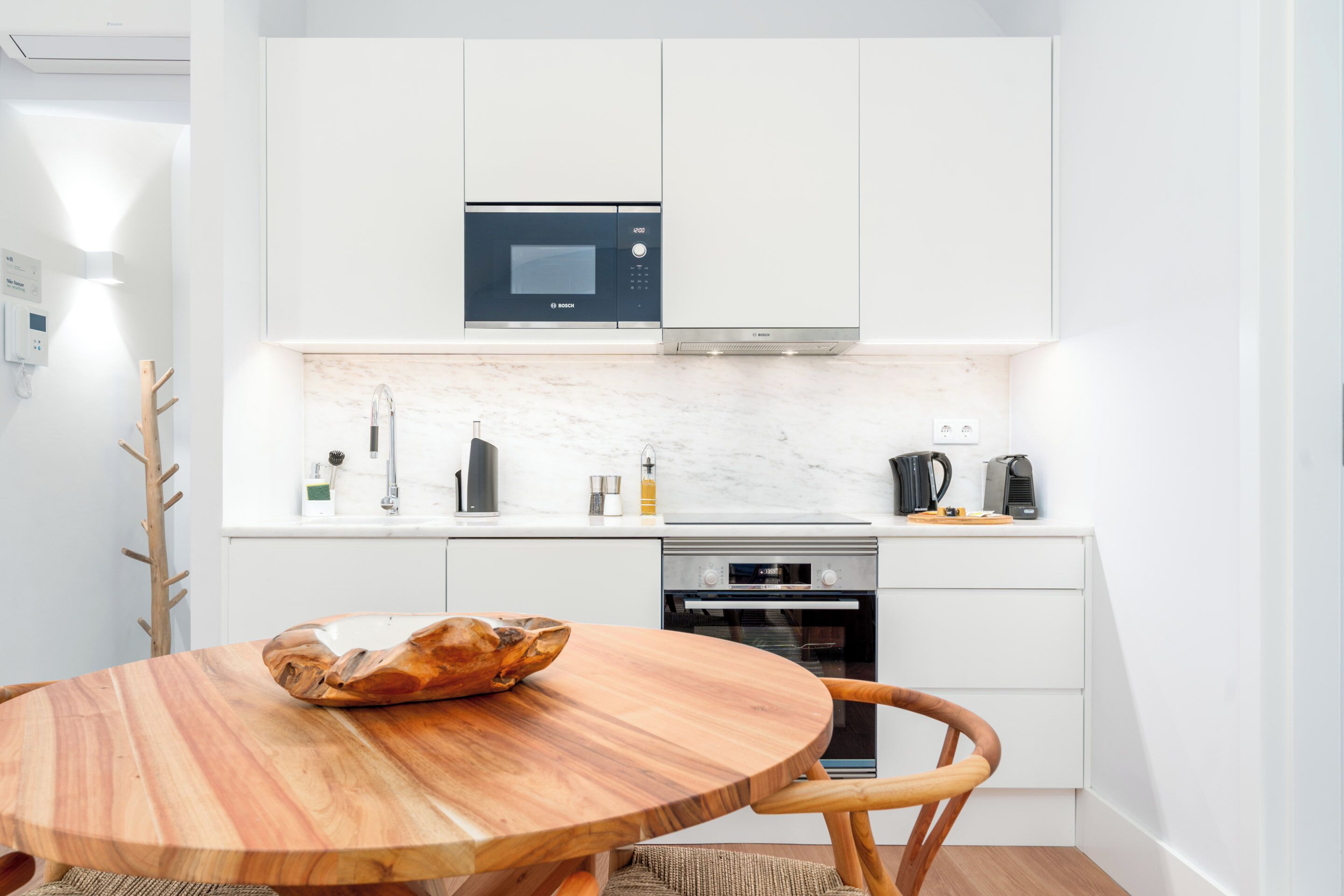 Una cocina moderna con armarios blancos, microondas integrado, estufa y una mesa redonda de madera con un bol.