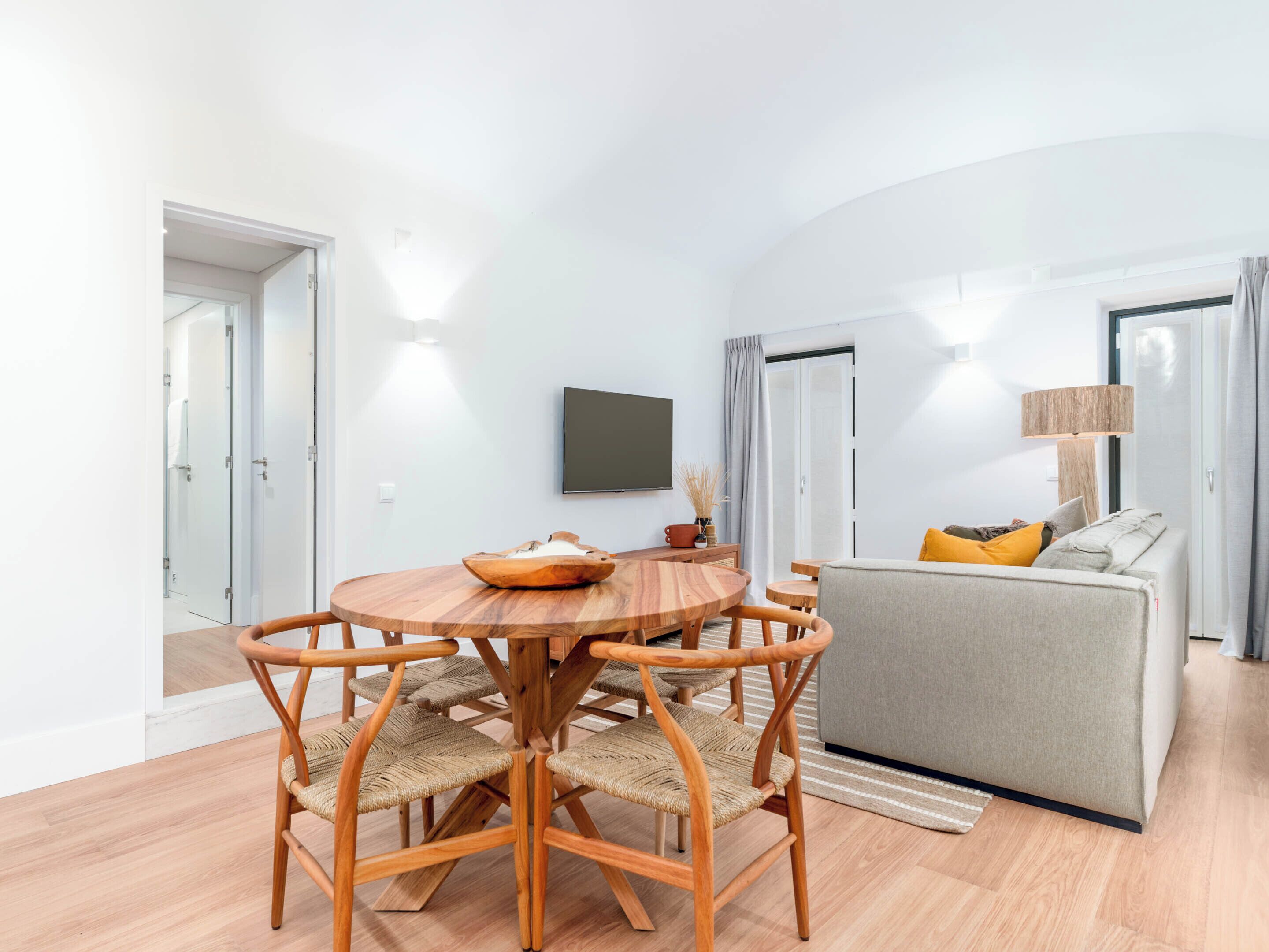 A modern living room with a round wooden dining table, four woven chairs, a grey sofa with yellow cushions, a flat-screen TV mounted on the wall, and a lamp on a side table.