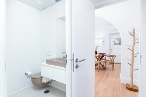 A modern bathroom with a white sink, toilet, and shower. There is a wooden coat rack and a dining area visible through an open door. - Limehome Evora Rua de Machede 61 (Évora)