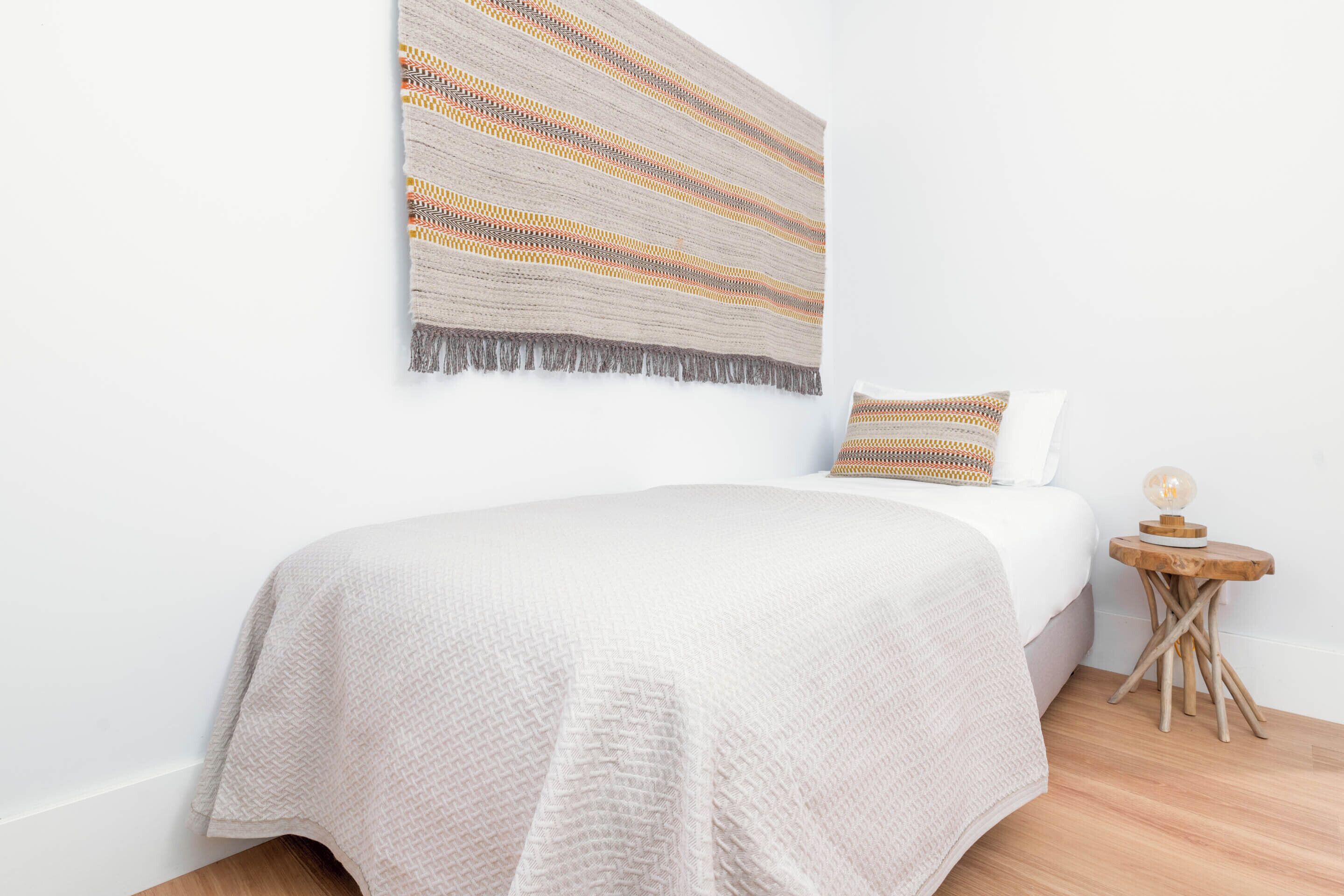 A neatly made bed with a white bedspread and a patterned throw blanket. There is a small wooden side table with a lamp beside the bed.