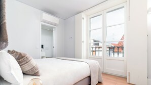 Un dormitorio con cama, mesita de noche con lámpara, ventana con vista a edificios y un equipo de aire acondicionado.