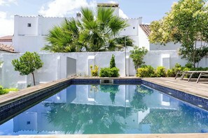 A clear swimming pool with a wooden deck, surrounded by white walls and greenery including palm trees and shrubs. - Limehome Evora Rua de Machede 61 (Évora)