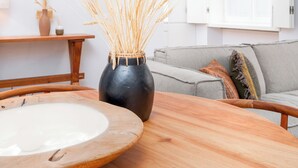 Una sala de estar moderna con una mesa redonda de madera, un jarrón oscuro con espigas de trigo, un sofá gris y armarios blancos.