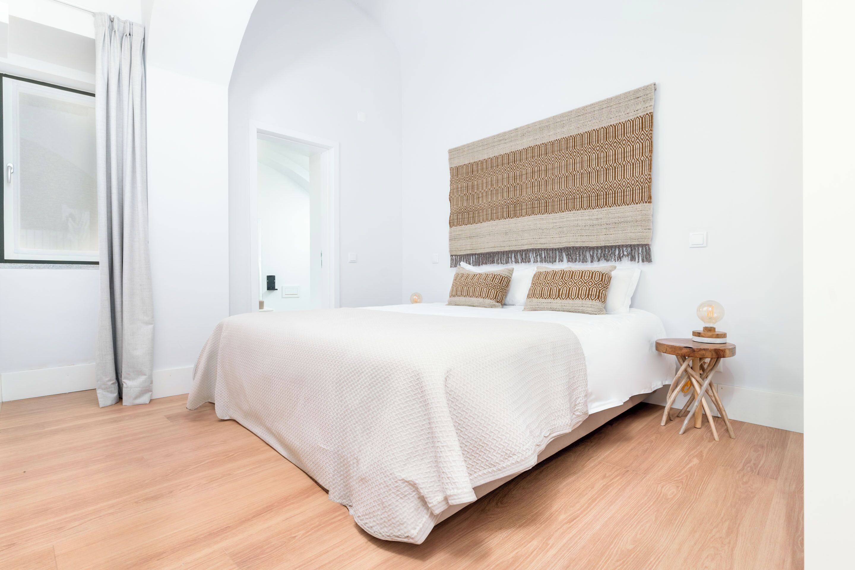 Une chambre à coucher avec un lit, une tête de lit ornée d’un motif, une table de chevet avec une lampe et une fenêtre avec des rideaux.