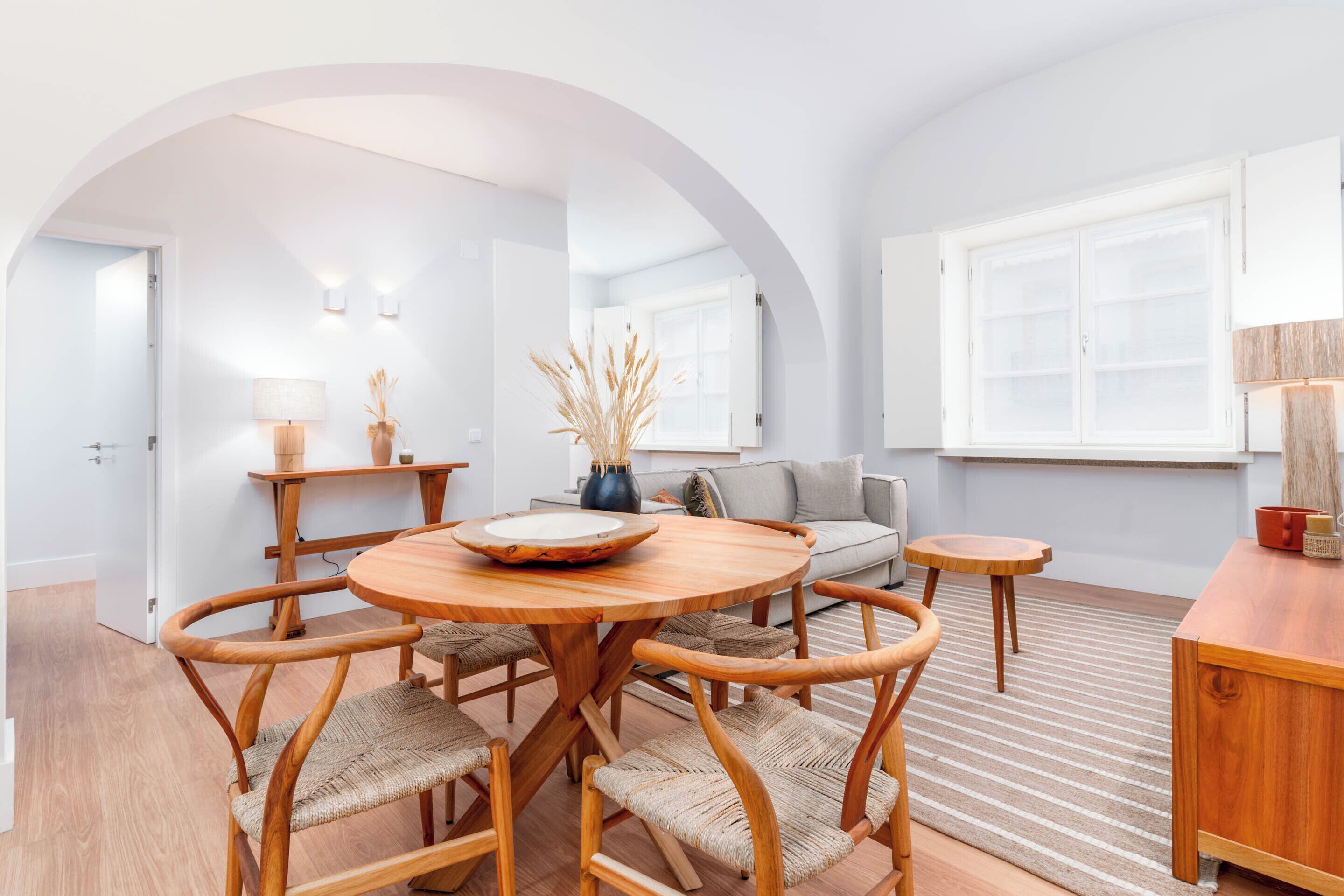A modern living room with a round wooden table, wicker chairs, a sofa, and a small wooden side table.