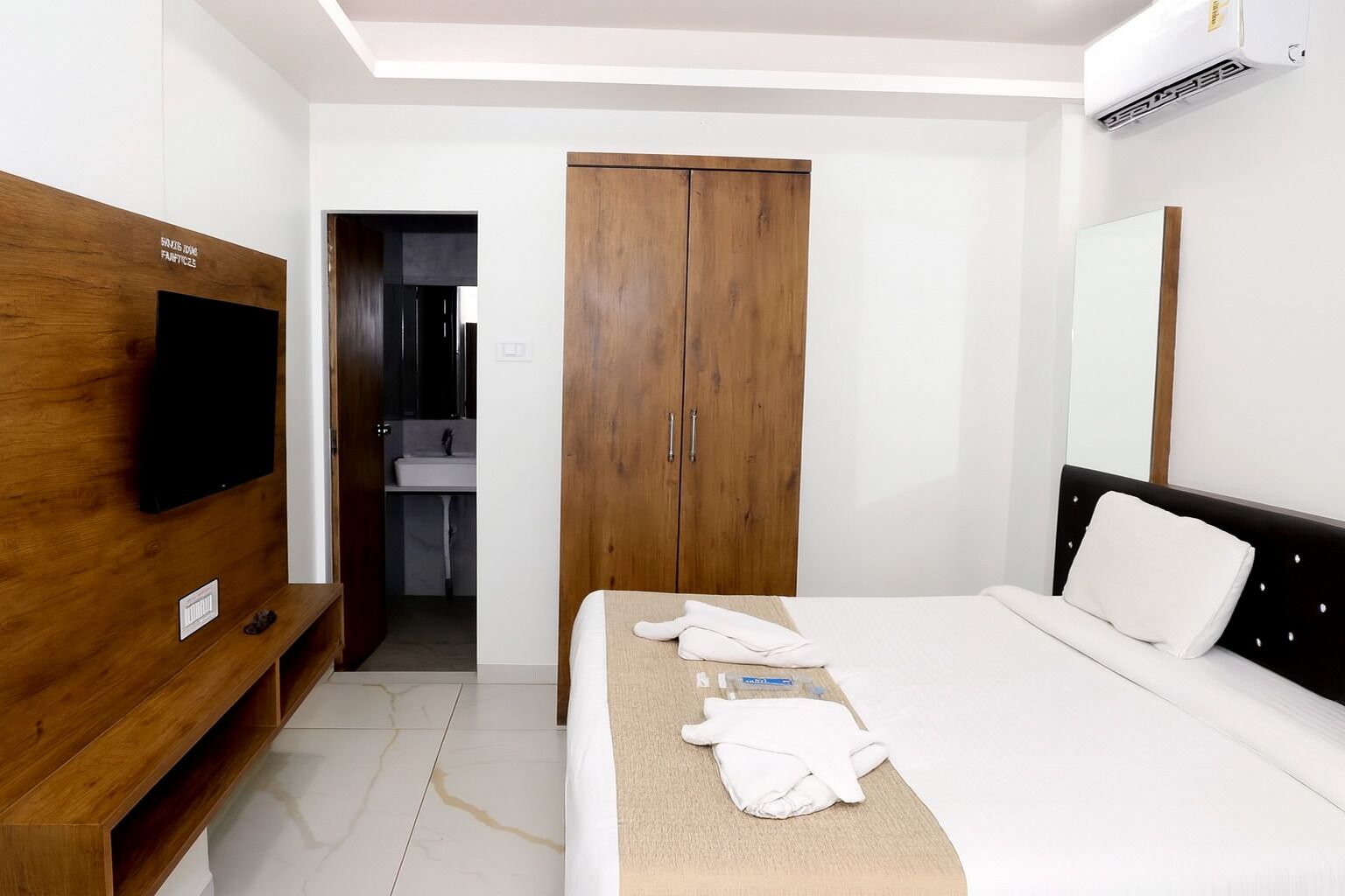 Quarto de hotel com cama, televisão fixada num painel de madeira, guarda-roupa e casa de banho visível através de uma porta aberta.