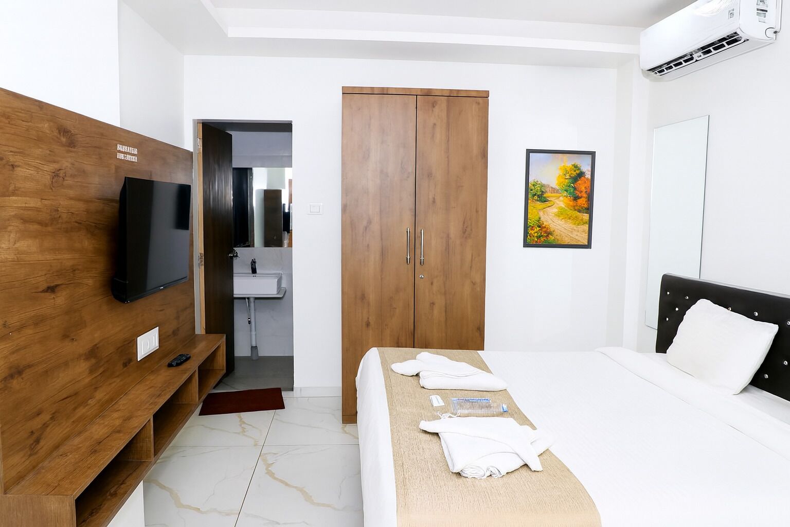 A modern hotel room with a wooden wardrobe, a flat-screen TV, a bed with white linens, a painting on the wall, and an air conditioning unit.