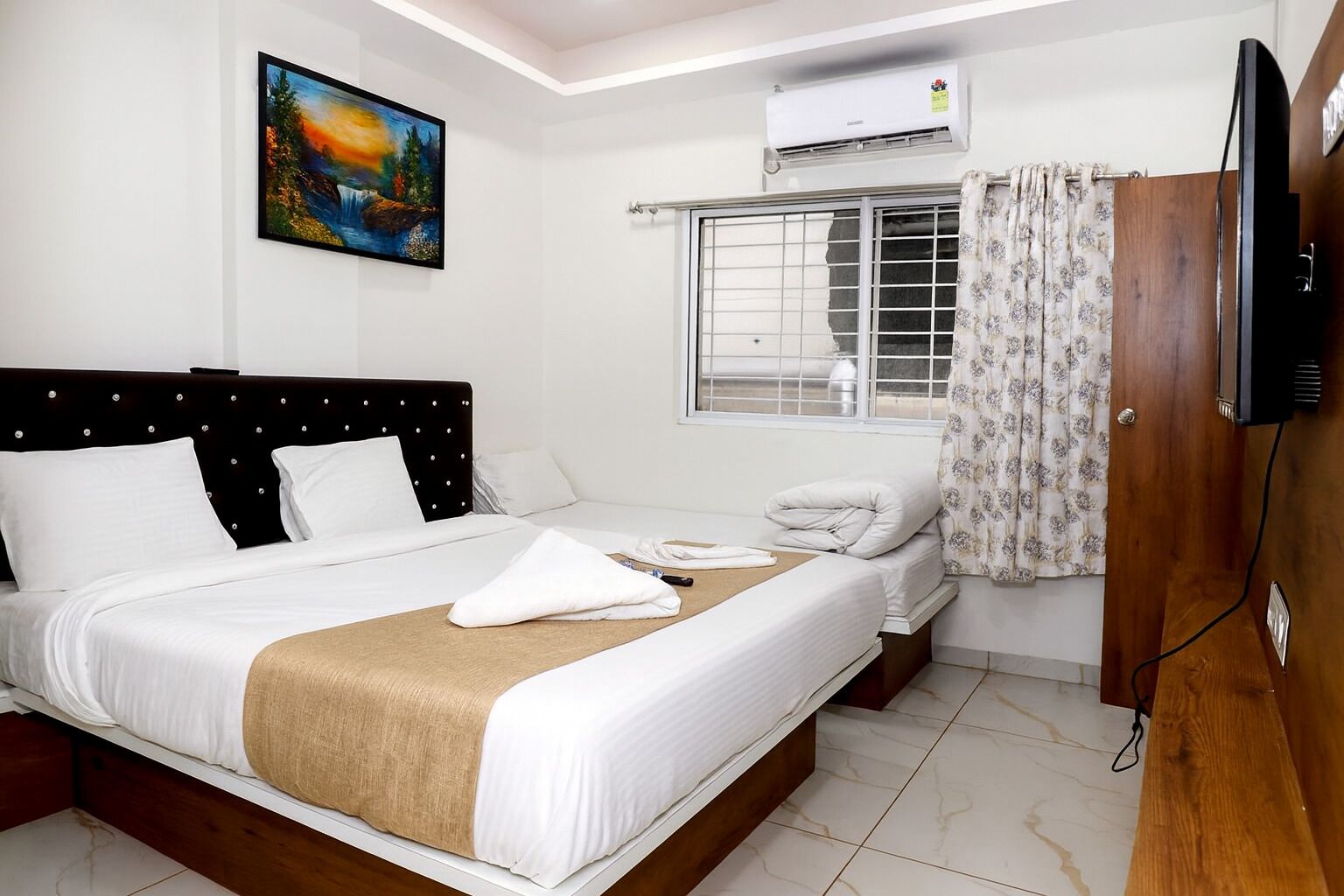 A hotel room with a bed, a painting, an air conditioner, a window with curtains, and a television.