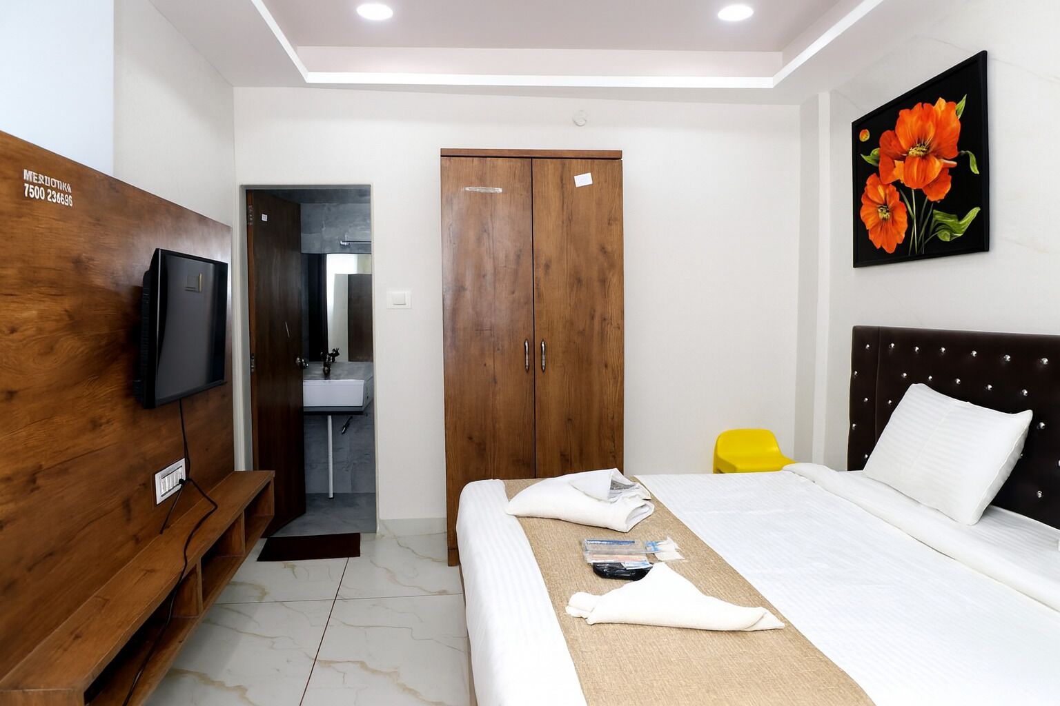 Un dormitorio con cama, un armario de madera, un televisor y un baño con lavamanos, además de un cuadro con flores.