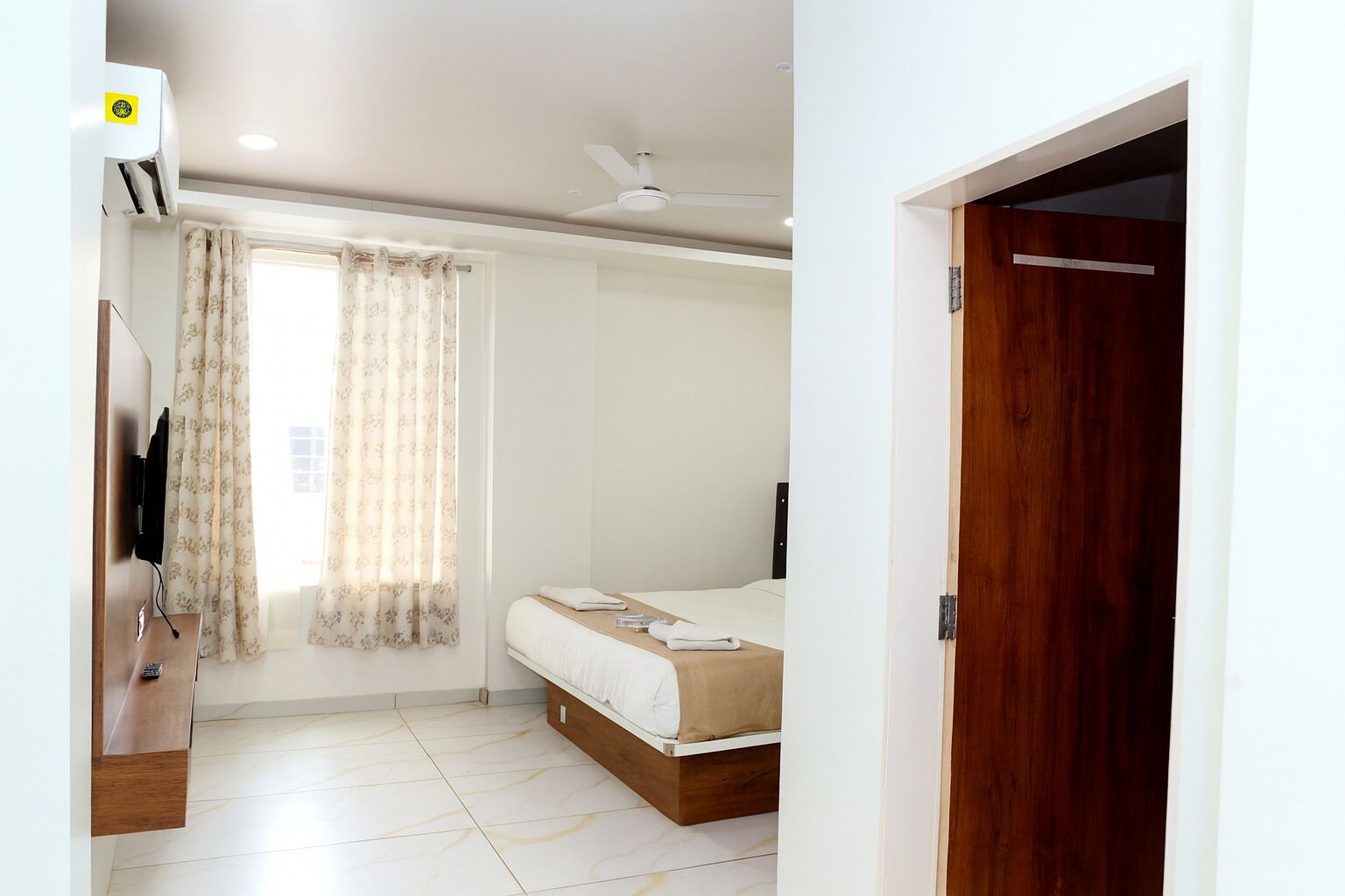 Habitació d'hotel amb un llit, una petita arca de fusta, un ventilador de sostre i una finestra amb cortines.