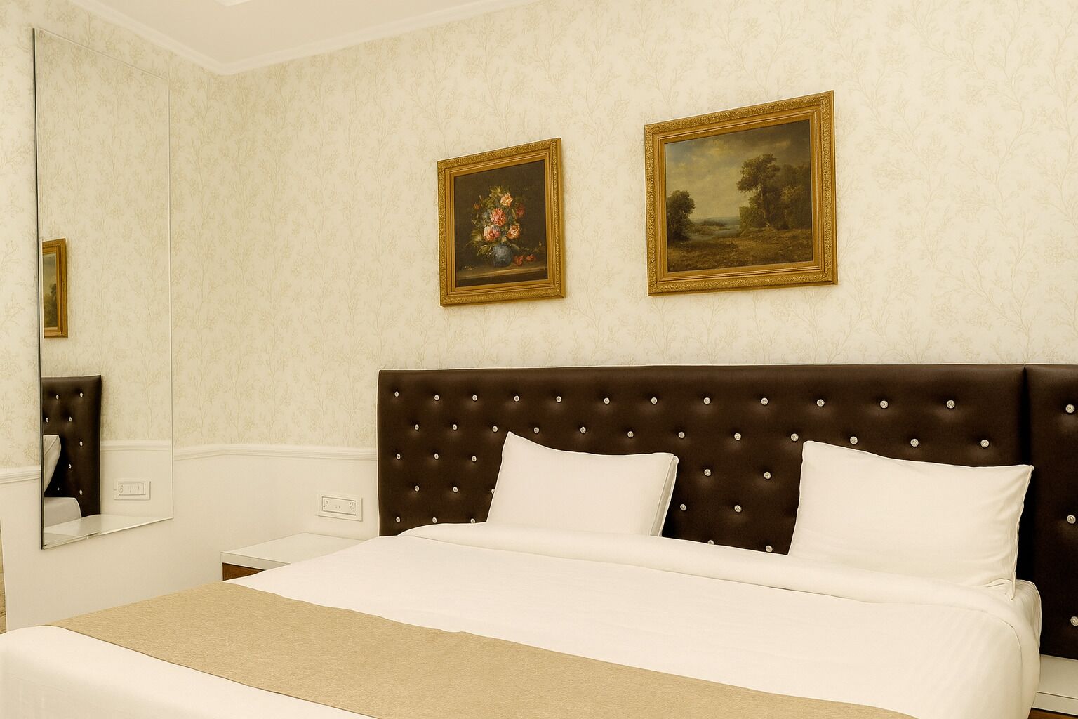 A hotel room with a large bed, a dark brown tufted headboard, two white pillows, a beige throw blanket, and two framed paintings on the wall.