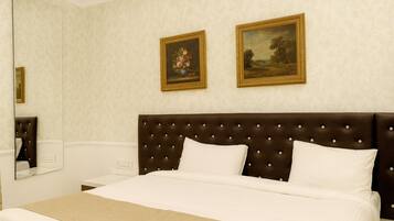 A hotel room with a large bed, a dark brown tufted headboard, two white pillows, a beige throw blanket, and two framed paintings on the wall.