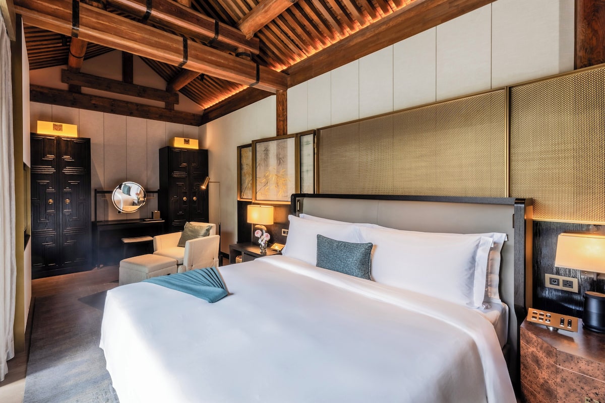 Habitación de hotel con una cama grande, vigas de techo de madera, un escritorio con silla, un espejo y obras de arte en las paredes.