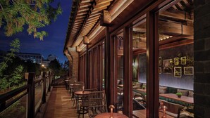 Un área de comedor al aire libre con mesas y sillas de madera, ventanas amplias y vistas a la ciudad al anochecer.