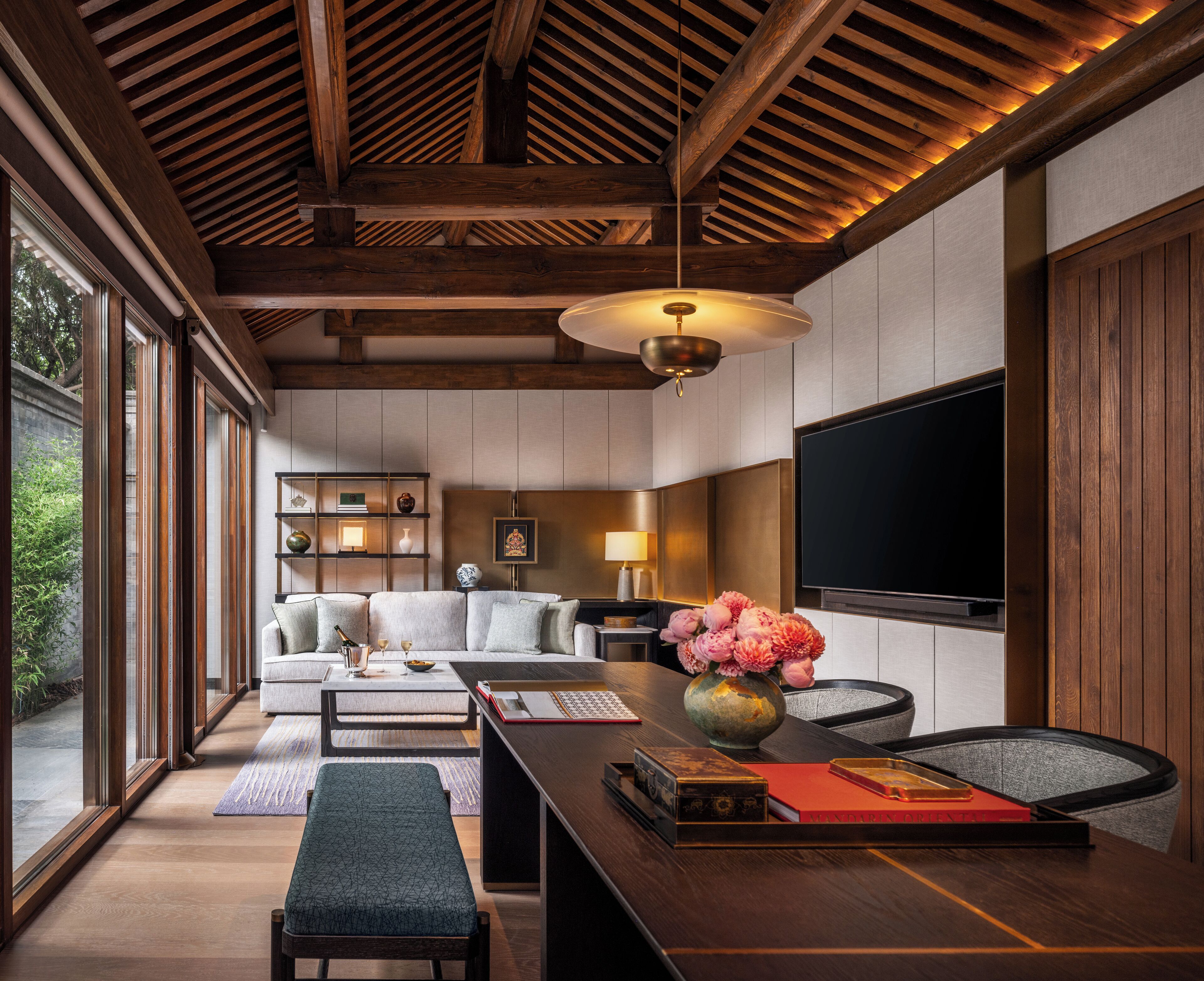 A modern living room with a wooden ceiling, a large flat-screen TV, a sofa, a coffee table, and a dining table with a vase of flowers.