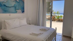 Ein Schlafzimmer mit Bett, weißer Bettwäsche, einem Gemälde an der Wand und Meerblick durch eine Schiebetür.
