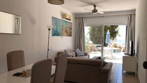 Ein modernes Wohnzimmer mit einer Couch, einem Esstisch und Stühlen. Ein Deckenventilator, ein Bild an der Wand und ein Blick ins Freie durch große Fenster sind ebenfalls vorhanden.