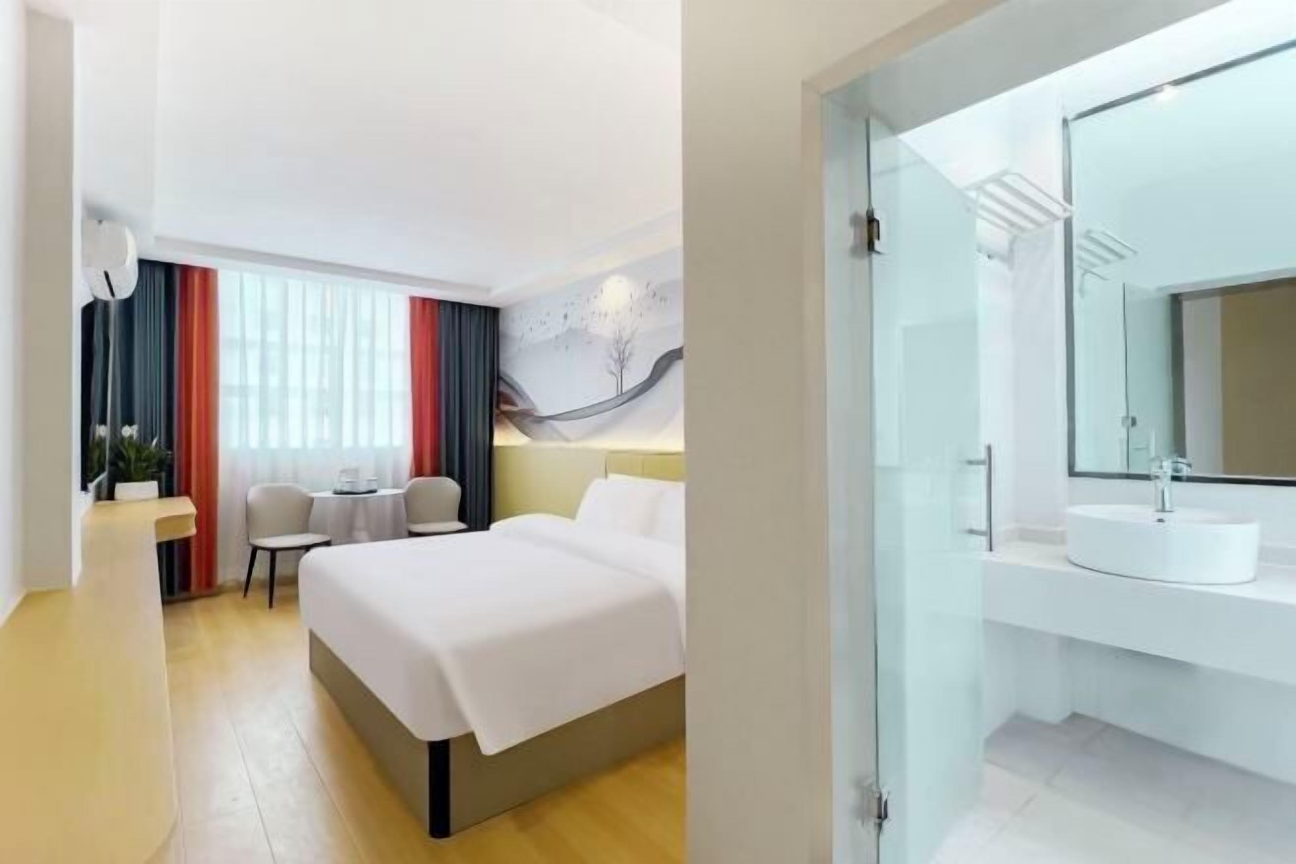A hotel room with a bed, bedside table, chair, and a glass-enclosed bathroom with a sink and mirror.