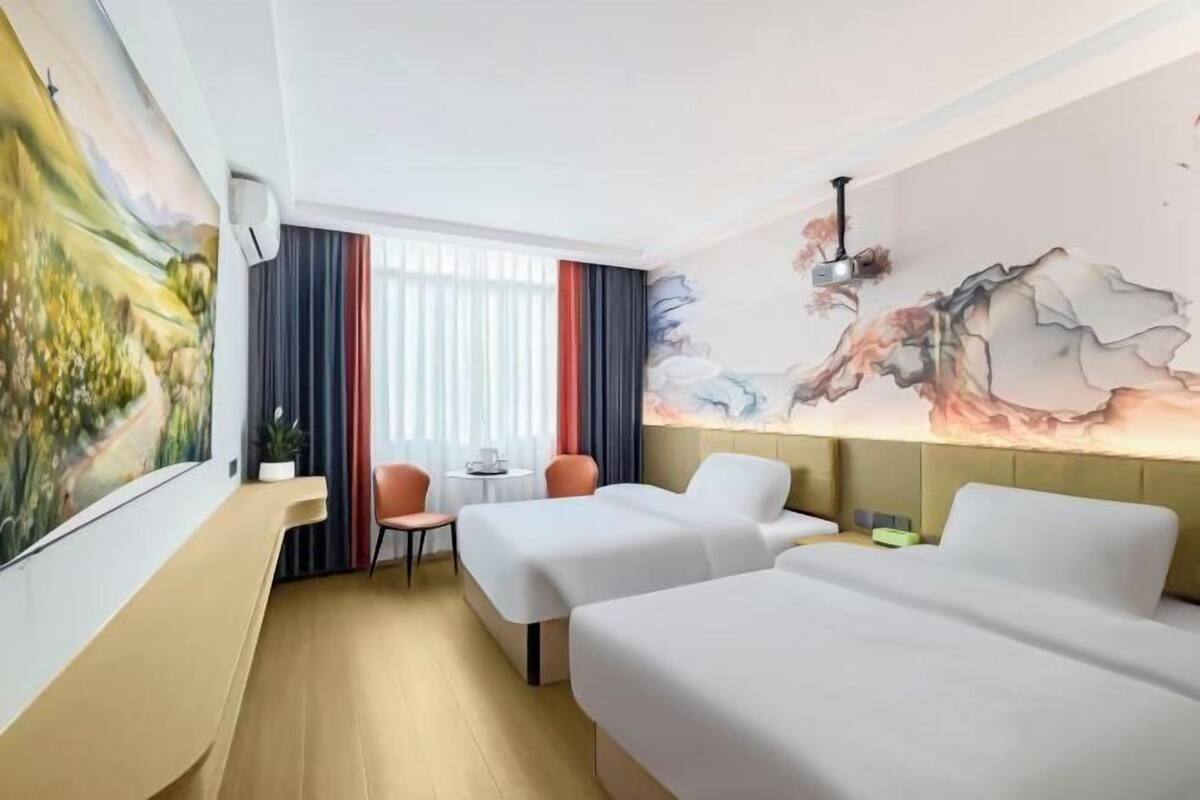 A hotel room with two beds, a desk, a chair, a window with curtains, and a wall with a painting.