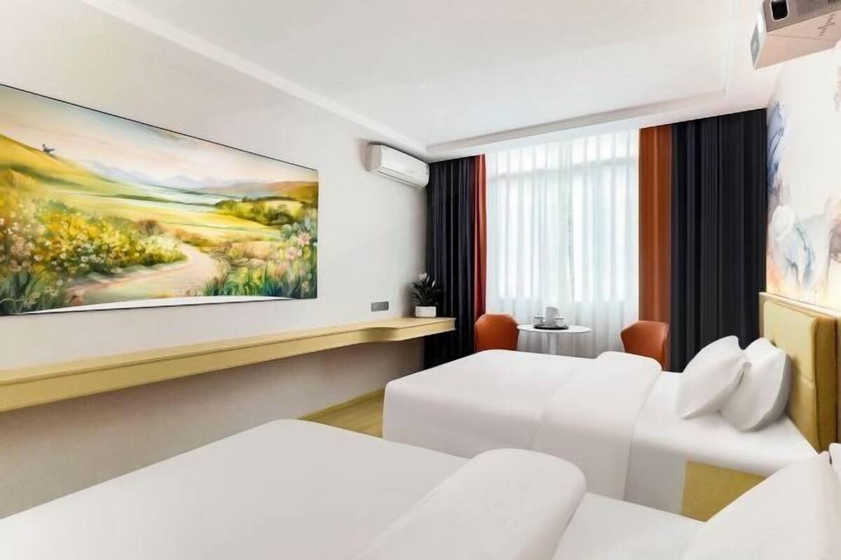 A hotel room with two beds, a painting of a landscape, a window with curtains, and a small table with a chair.