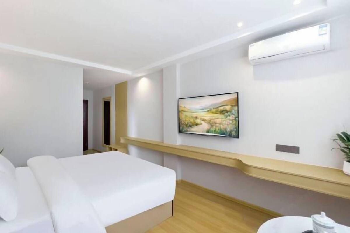 A hotel room with a bed, a mounted TV, a shelf, and an air conditioning unit.