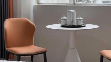 A hotel room with a single orange chair, a white table with a tea set, and a window with a view of buildings.