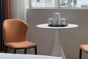 Quarto de hotel com uma única cadeira laranja, uma mesa branca com chá e janela com vista para prédios.