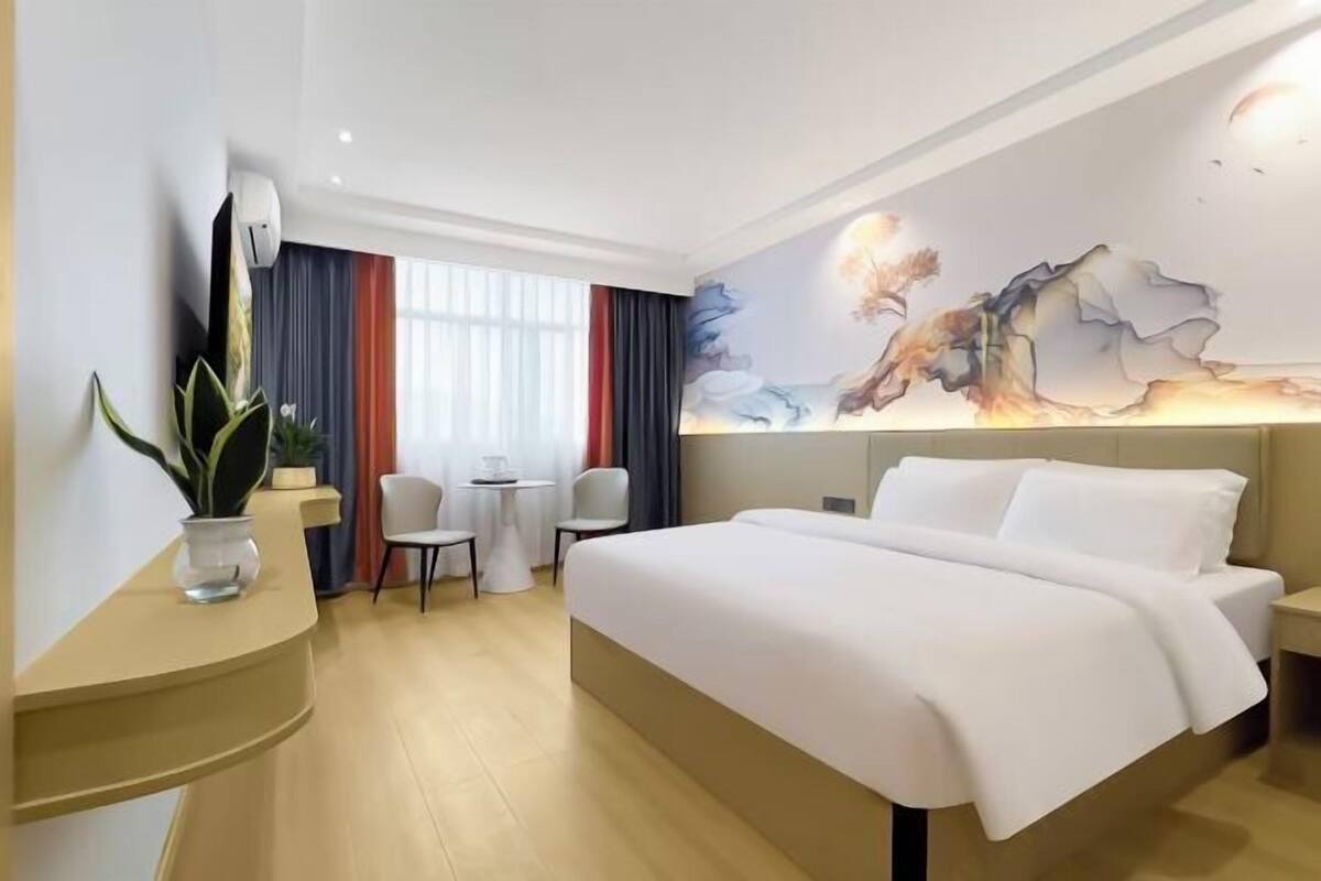 A hotel room with a bed, a desk, a chair, a small table, a potted plant, and a window with curtains.