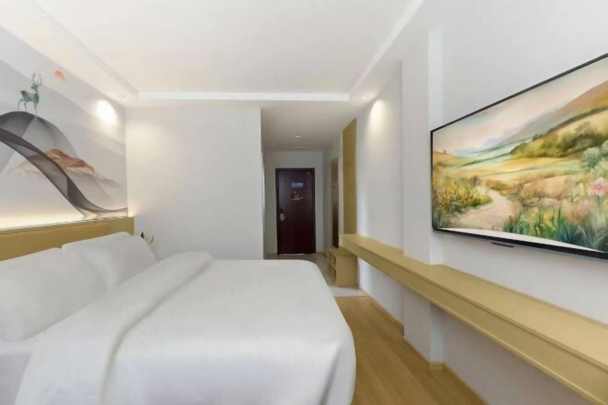 A hotel room with a bed, a painting on the wall, and a hallway leading to the exit.