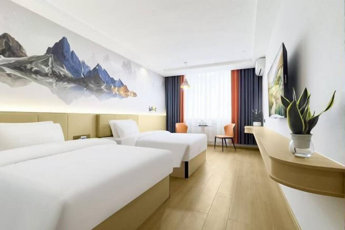 A hotel room with two beds, a desk, a chair, a large painting, and a plant.