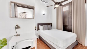 A bedroom with a bed, a desk, a mirror, a ceiling fan, and a window with curtains.