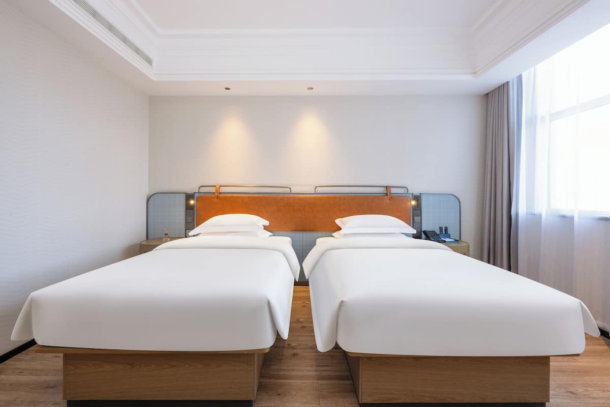 A hotel room with two single beds, a wooden headboard, a nightstand with a lamp, and a window with curtains.