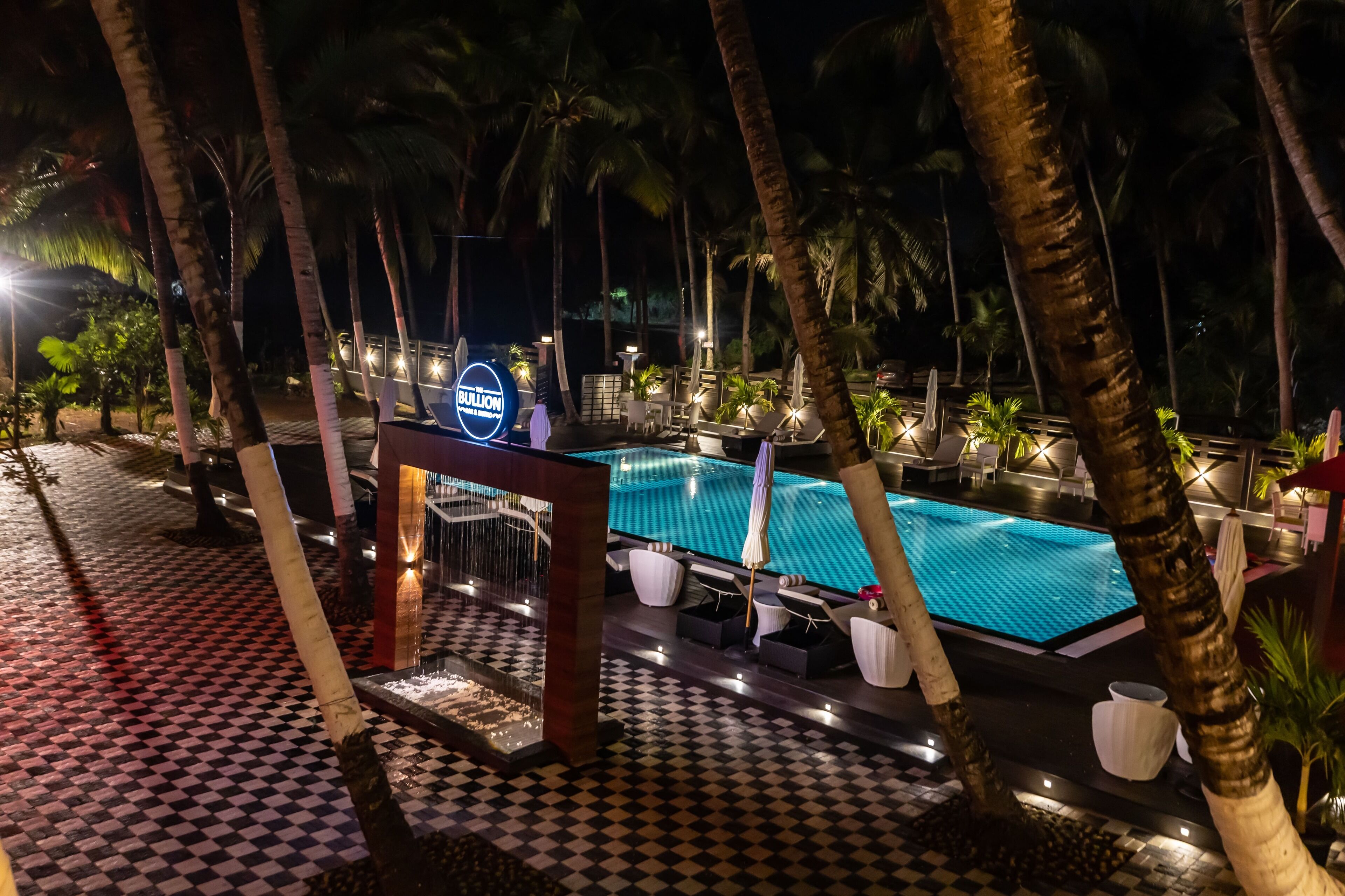 Un área de piscina nocturna con sillones reclinables y sombrillas, rodeada de palmeras y un estampado de cuadros en el suelo.