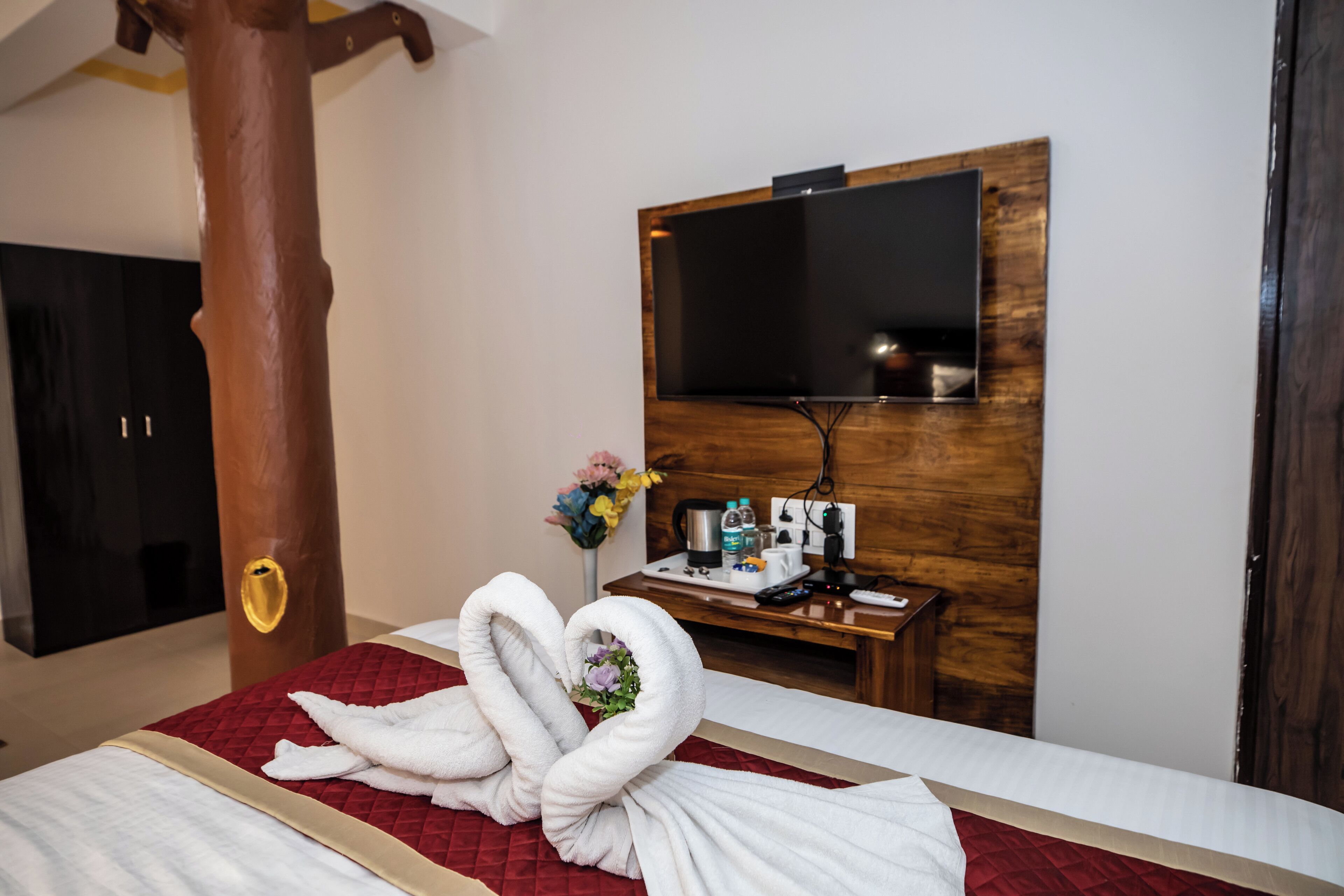 Hotelkamer met een bed, een houten pilaar, een flatscreen televisie aan de muur, een tafeltje met toiletartikelen en een bloemenboeket.