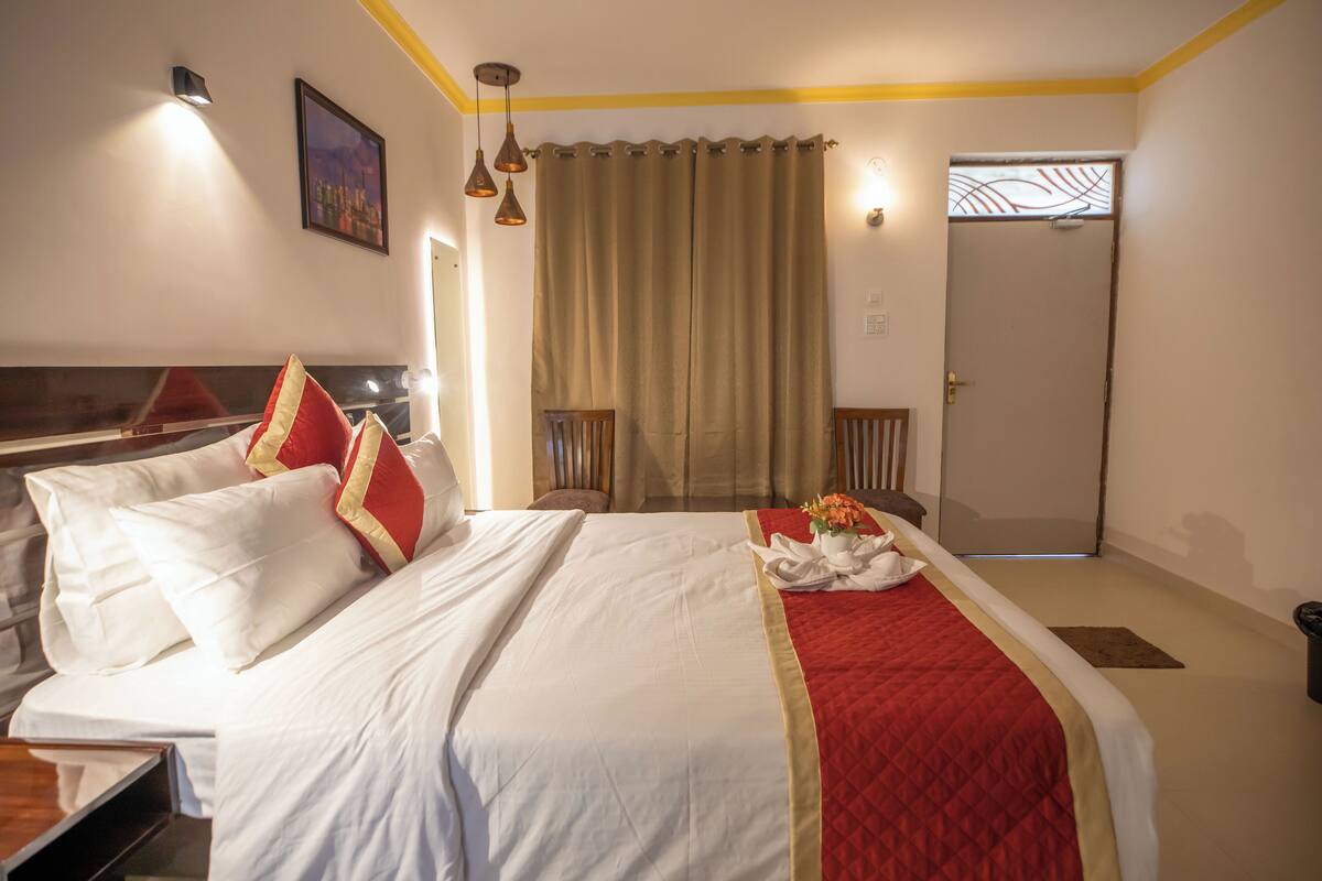 Una habitación de hotel con cama, escritorio, silla, ventana con cortinas, puerta y lámpara fijada a la pared.