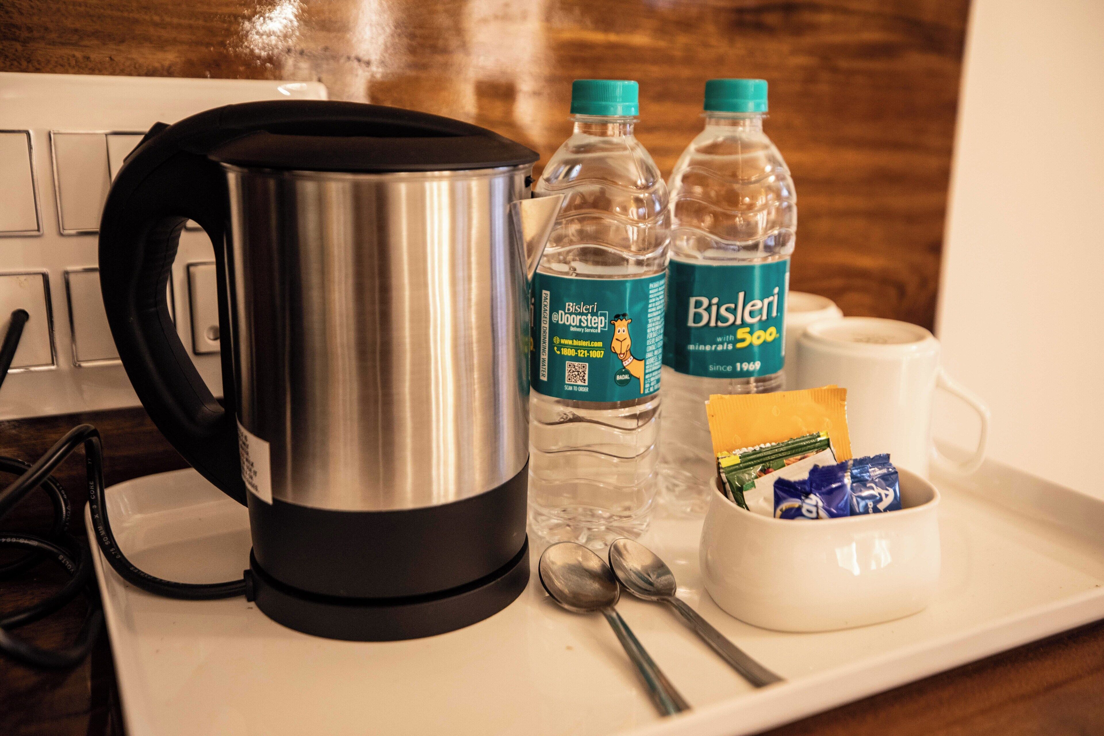 A kitchen counter with a stainless steel kettle, two Bisleri mineral water bottles, a white mug, a white cup, and a small white bowl containing snacks.